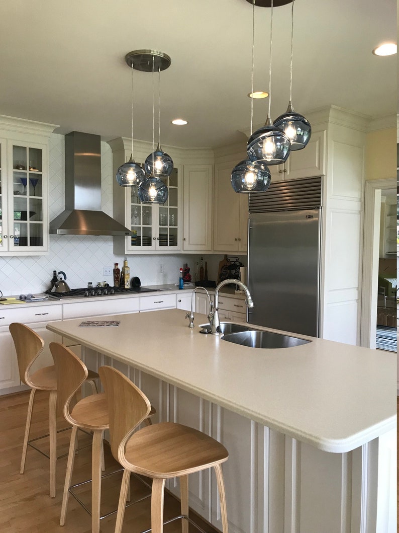 May include: A white kitchen island with a white countertop and a stainless steel sink. Three wooden bar stools with chrome legs are positioned around the island. A set of five blue glass pendant lights hang above the island.