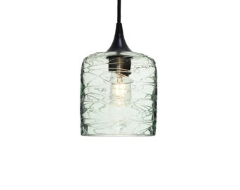 May include: A pendant light featuring a cylindrical, pale green glass shade with a textured, swirling surface pattern. The fixture includes a black metal cap and a black cord, with a single light bulb visible inside the glass housing.