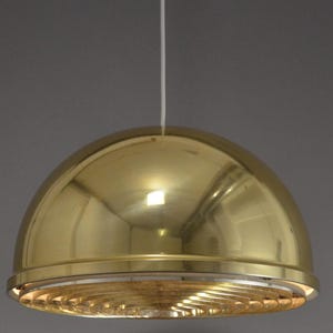 Puede incluir: Una lámpara colgante dorada en forma de cúpula. La luz tiene una superficie pulida y reflectante y un difusor circular con un patrón en espiral en la parte inferior. Cuelga de un cable blanco sobre un fondo gris neutro.