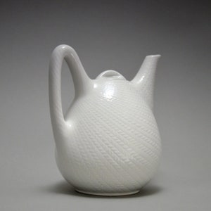 Puede incluir: Tetera de cerámica blanca con un patrón texturizado tejido. La tetera tiene un asa curva y un pico. La tapa tiene un pequeño pomo. La tetera es de color blanco liso y se asienta sobre un fondo neutro.