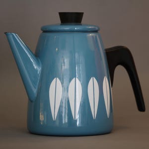 Könnte beinhalten: Eine blaue Emaille-Kaffeekanne im Vintage-Stil mit schwarzem Griff, Deckel und Knauf. Die Kanne hat weiße, stilisierte Blattmuster. Der Ausguss ist abgewinkelt und der Griff ist gebogen. Ein klassischer Küchenartikel.
