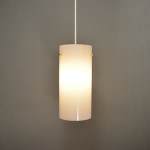 May include: A white cylindrical pendant light fixture with a brass top and bottom. The light is on and illuminating the room.
