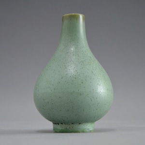 May include: A small, pale green ceramic vase with a speckled texture. The vase has a rounded body, a narrow neck, and a slightly flared base. The top rim is a darker shade of green.