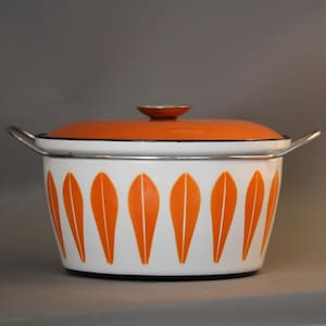 Könnte beinhalten: Ein weißer Emaille-Topf mit orangefarbenem Deckel und Griff. Der Topf hat ein sich wiederholendes Muster aus orangefarbenen blattartigen Formen. Der Deckel hat einen kleinen, runden Knopf. Der Topf hat einen silbernen Rand und Griffe.