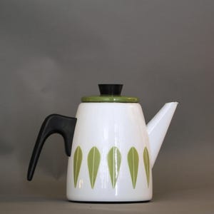Könnte beinhalten: Weiße Emaille-Teekanne mit schwarzem Griff und Deckel sowie grünem Deckel und Blattdesign. Die Teekanne hat einen Ausguss und wird wahrscheinlich zum Brühen von Tee oder Kaffee verwendet. Das Design ist modern.