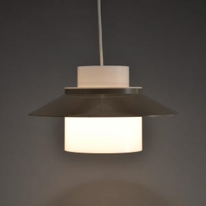 Puede incluir: Lámpara colgante moderna con una parte superior cilíndrica blanca y una pantalla gris en forma de platillo. La sección inferior es un cilindro blanco brillante. La luz cuelga de un cable blanco sobre un fondo gris.