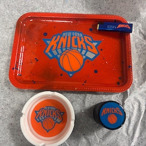 May include: A red rolling tray, ashtray, and container set featuring the New York Knicks logo. The tray has a basketball and team logo design. A blue lighter with the Knicks logo is also included.
