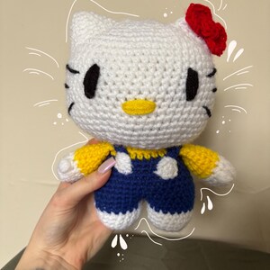 May include: A handmade crochet doll resembling a popular cartoon cat character. The doll has a white head with black eyes and a yellow nose. It wears a blue jumpsuit with white accents and a red bow. The arms are yellow and white.