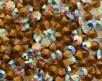 Puede incluir: Una imagen de primer plano de una gran cantidad de piedras de cristal doradas iridiscentes. Las piedras de cristal están esparcidas sobre una superficie blanca.