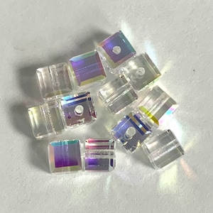 May include: A collection of clear, square-shaped beads with iridescent, rainbow-like color effects. Each bead has a small hole running through the center, suggesting they are designed for stringing. The beads are scattered on a light-colored surface.