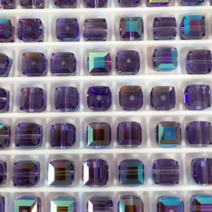 May include: A close-up view of a tray of square-shaped glass beads. The beads are a deep purple color with iridescent highlights. The beads are arranged in rows and columns on a white tray.