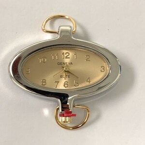 May include: A silver and gold oval-shaped pocket watch with a gold-coloured face and black hands. The watch face has black numbers and the words "GENEVA" and "ELITE" printed on it.