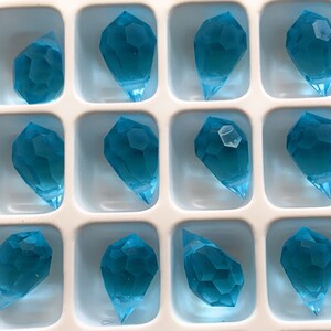 K&ouml;nnte beinhalten: Ein Tablett mit zw&ouml;lf facettierten, tropfenf&ouml;rmigen, aquamarinblauen Kristallanh&auml;ngern. Jeder Anh&auml;nger hat eine spitze Unterseite und befindet sich in einem quadratischen Fach. Die Kristalle haben ein geometrisches Design.