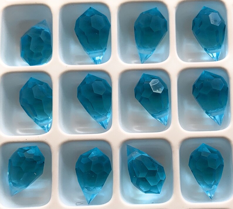 K&ouml;nnte beinhalten: Ein Tablett mit zw&ouml;lf facettierten, tropfenf&ouml;rmigen, aquamarinblauen Kristallanh&auml;ngern. Jeder Anh&auml;nger hat eine spitze Unterseite und befindet sich in einem quadratischen Fach. Die Kristalle haben ein geometrisches Design.