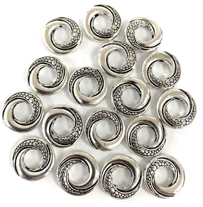 May include: A collection of 15 silver-toned metal beads with a swirl design. Each bead has a circular shape with a hole in the center. The beads are perfect for jewelry making or crafting.