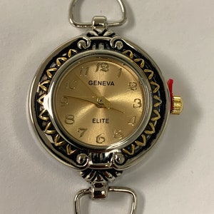 May include: A silver and gold tone wristwatch with a round face and a decorative border. The face is gold with black hands and black Roman numeral markings. The watch face says "GENEVA" and "ELITE".