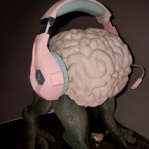 May include: A pink and light blue gaming headset rests on a light pink brain sculpture. The sculpture is mounted on a dark green, reptilian-like body with clawed feet. The headset has the brand name "OZIBO" printed on the side.