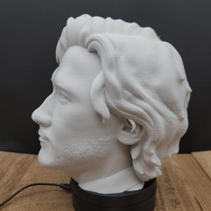 May include: A white 3D-printed bust of a man's head and shoulders, displayed on a black rotating base. The bust has detailed facial features and wavy hair. A business card with the text "Ms. Rabbit Trinkets" is in front.