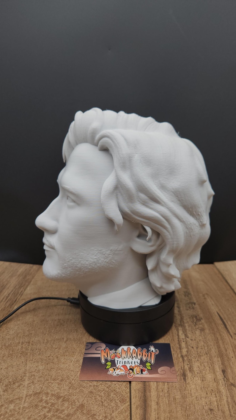 May include: A white 3D-printed bust of a man's head and shoulders, displayed on a black rotating base. The bust has detailed facial features and wavy hair. A business card with the text "Ms. Rabbit Trinkets" is in front.