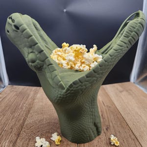 Peut inclure: Un bol vert en forme de main texturée rempli de pop-corn. Le bol est posé sur une surface en bois, avec du pop-corn éparpillé et une carte de visite à proximité. Le bol est conçu comme une griffe de reptile tenant le pop-corn.