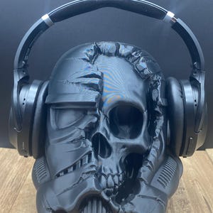 May include: Black skull-shaped headphone stand with a detailed design, holding black over-ear headphones. The stand has a textured surface and is placed on a wooden surface. The overall theme is a unique and decorative headphone holder.
