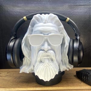 Pode incluir: Um busto branco de um homem barbudo usando óculos de sol brancos, com fones de ouvido pretos na cabeça. O busto está em uma base preta, e um mouse de computador preto é visível.