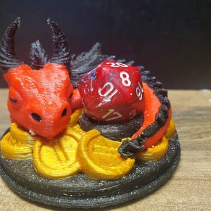 May include: A red and black 3D printed dragon figurine with a red 20-sided die resting on its back. The dragon is curled up on a bed of gold coins.