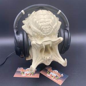May include: A cream-colored Predator head sculpture with a wide open mouth, holding a pair of black headphones. Two small business cards are at the base of the sculpture. The background is a dark gray.