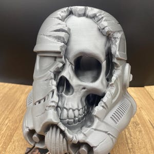May include: A gray, 3D-printed skull sculpture, with a Stormtrooper helmet design partially peeled away to reveal the skull beneath. The sculpture is on a wooden surface, with a logo visible at the bottom.