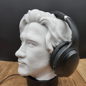 May include: A white bust of a man with a beard and headphones. The bust is on a black base, and the headphones are black. The bust is made of a light-colored material, and the headphones are resting on the bust's head.