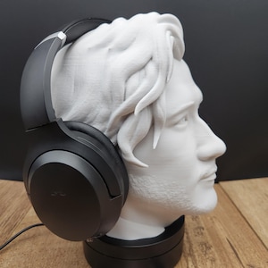 May include: A white bust of a man with black headphones resting on the head. The bust is on a black base, and the headphones are black. The background is black and the surface is wood. The logo "MoshRabbit Trinkets" is visible.