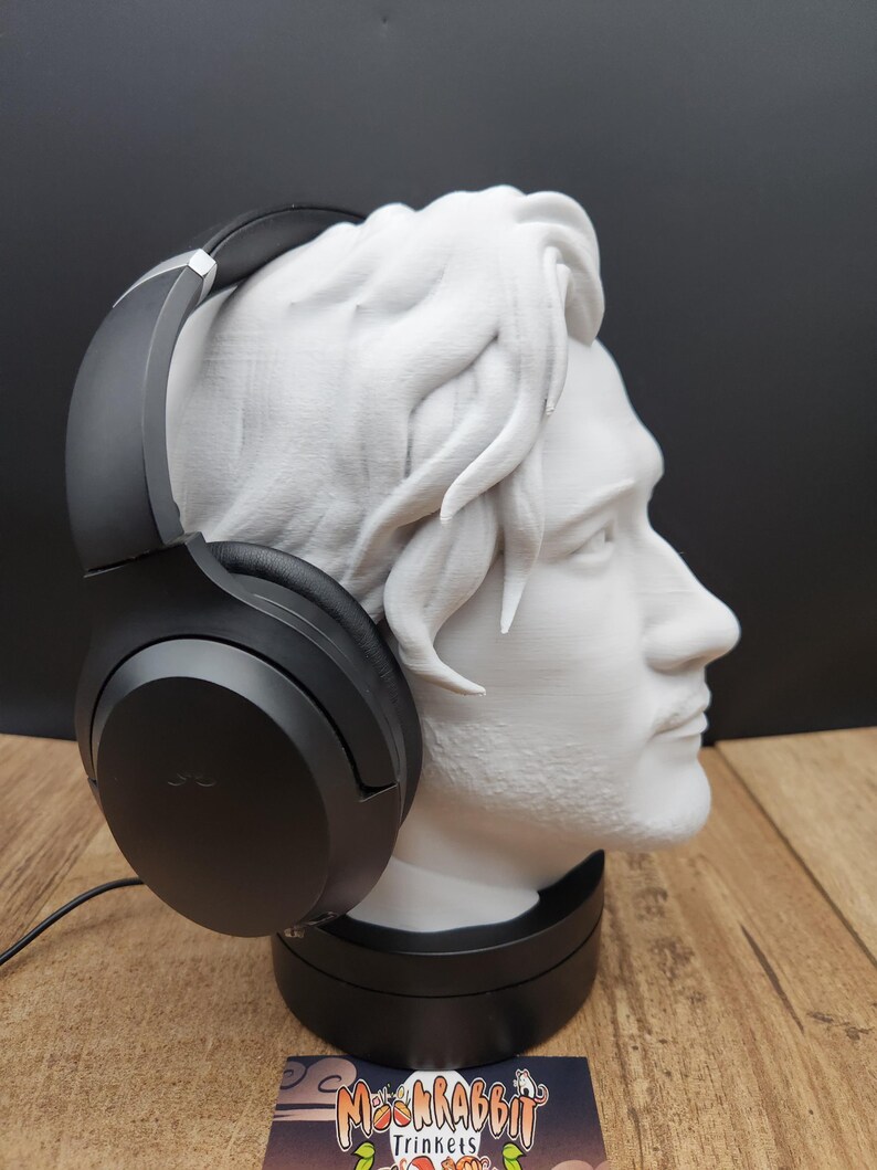 May include: A white bust of a man with black headphones resting on the head. The bust is on a black base, and the headphones are black. The background is black and the surface is wood. The logo "MoshRabbit Trinkets" is visible.
