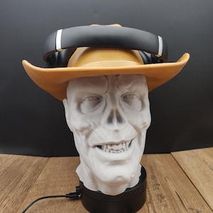 May include: A white skull-shaped head wearing a tan cowboy hat and black headphones. The skull is on a black base with a cord. The logo "Moonrabbit Trinkets" is visible at the bottom.