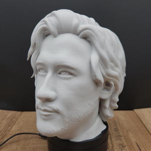 May include: A white 3D-printed bust of a person with wavy hair and a beard, mounted on a black base. The bust is a detailed sculpture with a realistic facial expression. A business card with the logo "Moonrabbit Trinkets" is in the foreground.