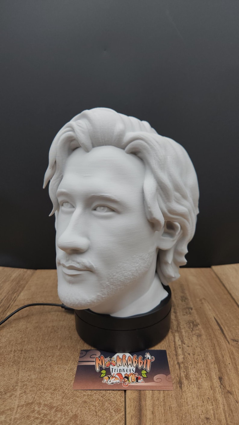 May include: A white 3D-printed bust of a person with wavy hair and a beard, mounted on a black base. The bust is a detailed sculpture with a realistic facial expression. A business card with the logo "Moonrabbit Trinkets" is in the foreground.