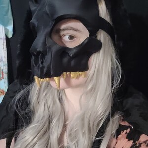 K&ouml;nnte beinhalten: Eine schwarze Totenkopfmaske mit goldenen Z&auml;hnen. Die Maske hat ein katzenartiges Design mit spitzen Ohren und einem breiten Mund. Die Maske wird von einer Person mit langen, silbernen Haaren getragen.