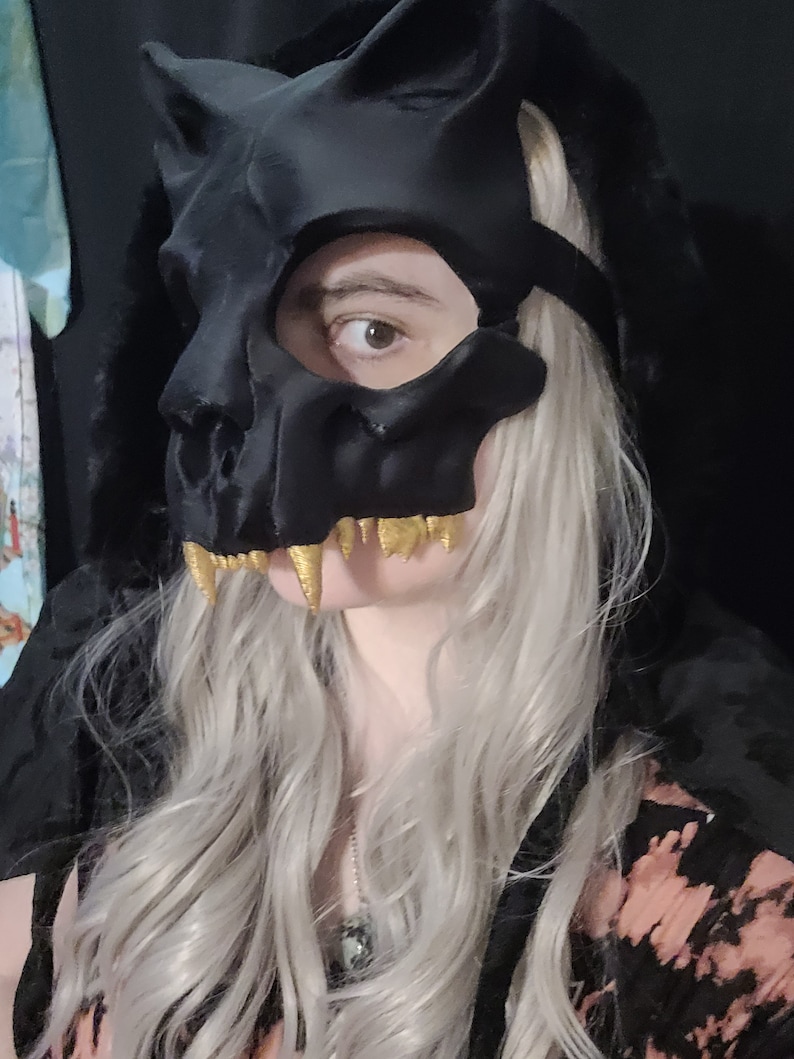 K&ouml;nnte beinhalten: Eine schwarze Totenkopfmaske mit goldenen Z&auml;hnen. Die Maske hat ein katzenartiges Design mit spitzen Ohren und einem breiten Mund. Die Maske wird von einer Person mit langen, silbernen Haaren getragen.