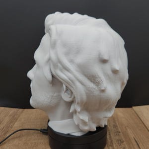May include: A white bust of a man's head, viewed in profile, with detailed hair and facial features. The bust sits on a black rotating base. A business card with a logo is visible in the foreground.