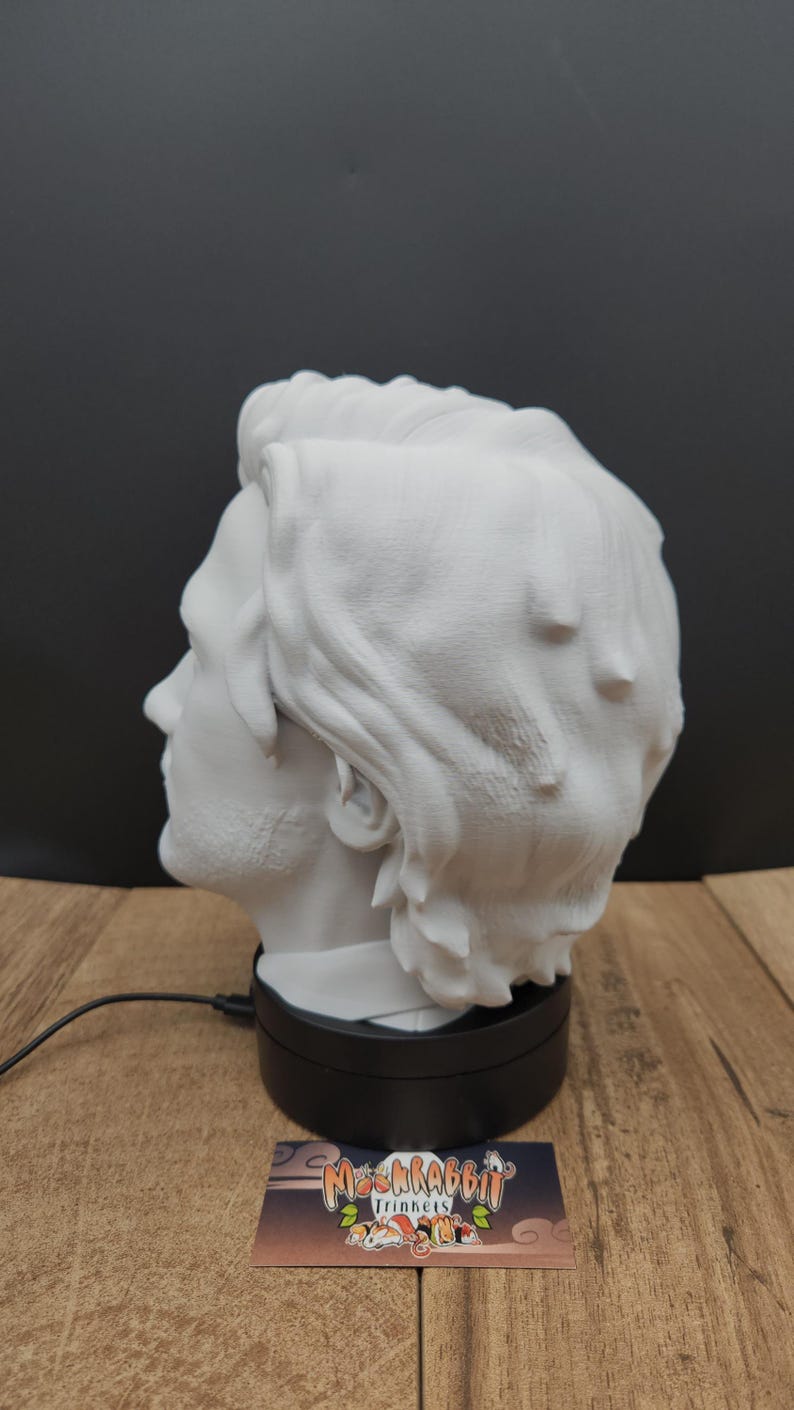 May include: A white bust of a man's head, viewed in profile, with detailed hair and facial features. The bust sits on a black rotating base. A business card with a logo is visible in the foreground.