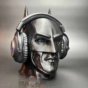 May include: A black Batman-themed headphone stand with a detailed face and cowl design. The stand features two pointed ears and holds a pair of black headphones. The stand is made of a textured material and has a metallic sheen.