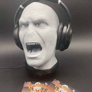 May include: A grey bust of a character with headphones, featuring an open mouth and detailed facial features. The headphones are black with silver accents. Two business cards with "MossRabbit Trinkets" are at the bottom.