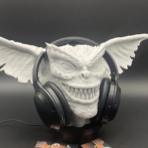 May include: A white, sculpted Gremlin head with large ears and wings, holding black headphones. Two business cards with the text "Mrs. Rabbit Trinkets" are at the base. The Gremlin has a wide grin and detailed facial features.