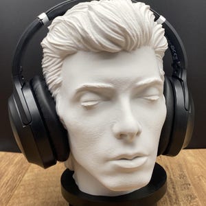 May include: A white bust of a man with closed eyes, wearing black headphones. The bust is on a black rotating base. The background is black and the surface is wood. The bust is a decorative item.