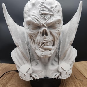 May include: A detailed, light gray bust of a skeletal figure with large, wing-like protrusions. The figure has a stitched head and two skull-shaped accents. The bust is mounted on a black base, with a logo visible at the bottom.