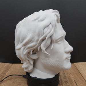 May include: A white 3D-printed bust of a man's head, showcasing detailed facial features and wavy hair. The bust is mounted on a black rotating base. The background is black, and the base sits on a wooden surface.