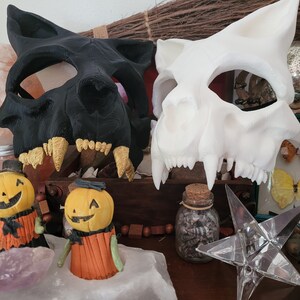 K&ouml;nnte beinhalten: Zwei Halloween-Masken, eine schwarze und eine wei&szlig;e, werden auf einem Tisch mit verschiedenen Dekorationsartikeln ausgestellt. Die Masken sind beide Tiersch&auml;del mit scharfen Z&auml;hnen. Die schwarze Maske hat goldene Z&auml;hne, w&auml;hrend die wei&szlig;e Maske wei&szlig;e Z&auml;hne hat. Beide Masken sind aus hartem Kunststoff.