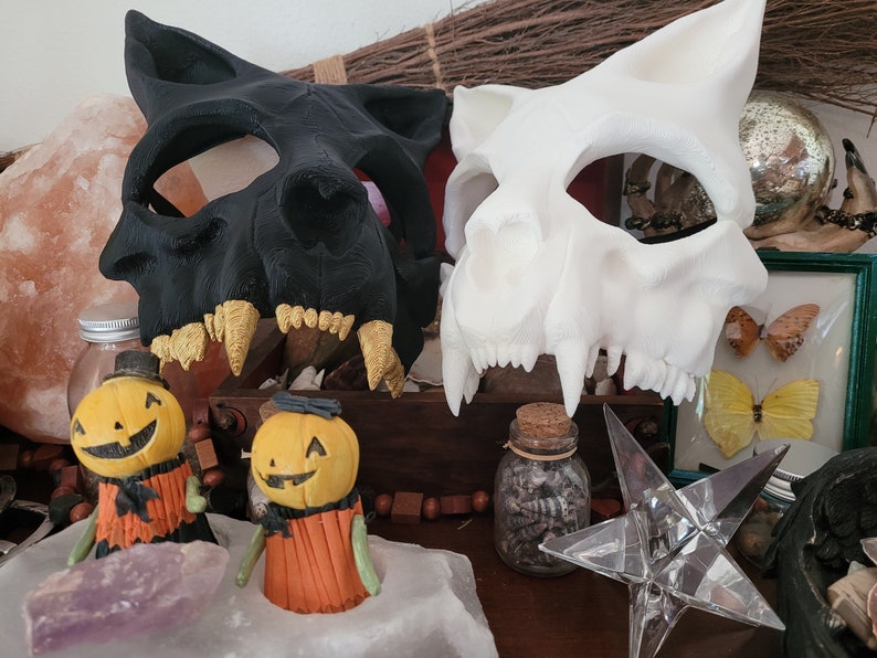 K&ouml;nnte beinhalten: Zwei Halloween-Masken, eine schwarze und eine wei&szlig;e, werden auf einem Tisch mit verschiedenen Dekorationsartikeln ausgestellt. Die Masken sind beide Tiersch&auml;del mit scharfen Z&auml;hnen. Die schwarze Maske hat goldene Z&auml;hne, w&auml;hrend die wei&szlig;e Maske wei&szlig;e Z&auml;hne hat. Beide Masken sind aus hartem Kunststoff.