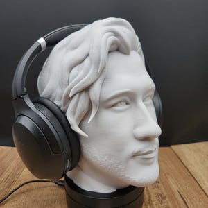 May include: A white bust of a man with headphones resting on the head. The bust is on a black base. The headphones are black with silver accents. The bust has detailed hair and facial features. The background is black and the surface is wood.