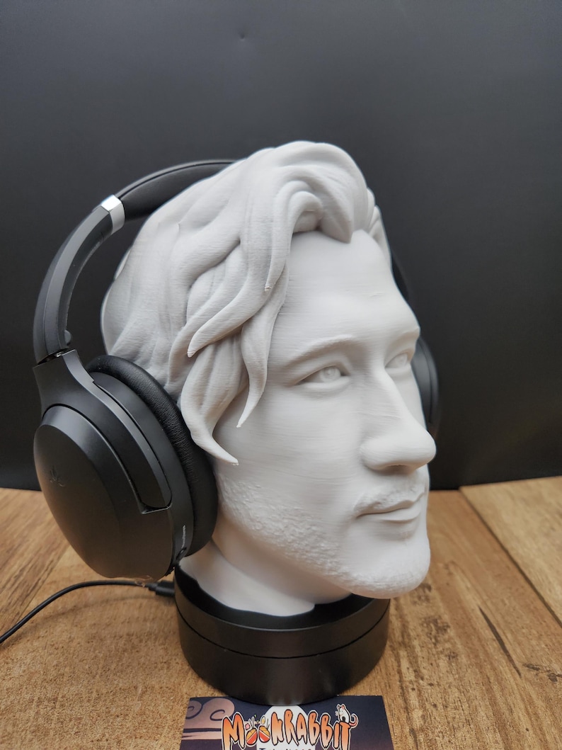 May include: A white bust of a man with headphones resting on the head. The bust is on a black base. The headphones are black with silver accents. The bust has detailed hair and facial features. The background is black and the surface is wood.