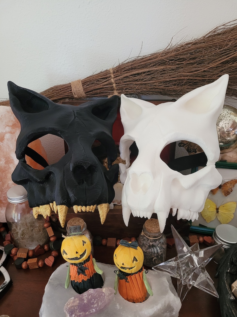 K&ouml;nnte beinhalten: Zwei Halloween-Masken, eine schwarze und eine wei&szlig;e, sind in Form von Tiersch&auml;deln mit Z&auml;hnen gestaltet. Die Masken liegen auf einem Tisch mit anderen Halloween-Dekorationen.